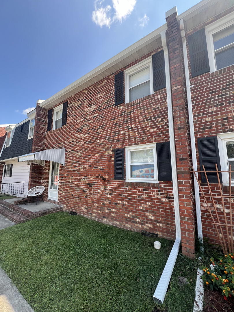 brick house with white gutters and black shutters green grass porch with a white chair and landscaping along the edge